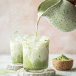 A hand pours milk into a glass of iced matcha latte, creating a swirling green and white effect, with another glass and a bowl of matcha powder in the background.